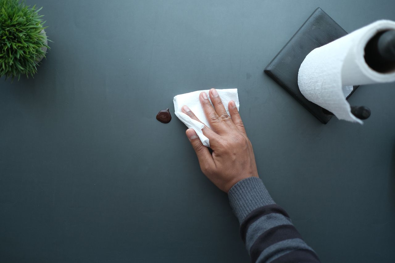 Une main essuie une surface foncée avec un mouchoir près d’un rouleau de papier et d’une plante.